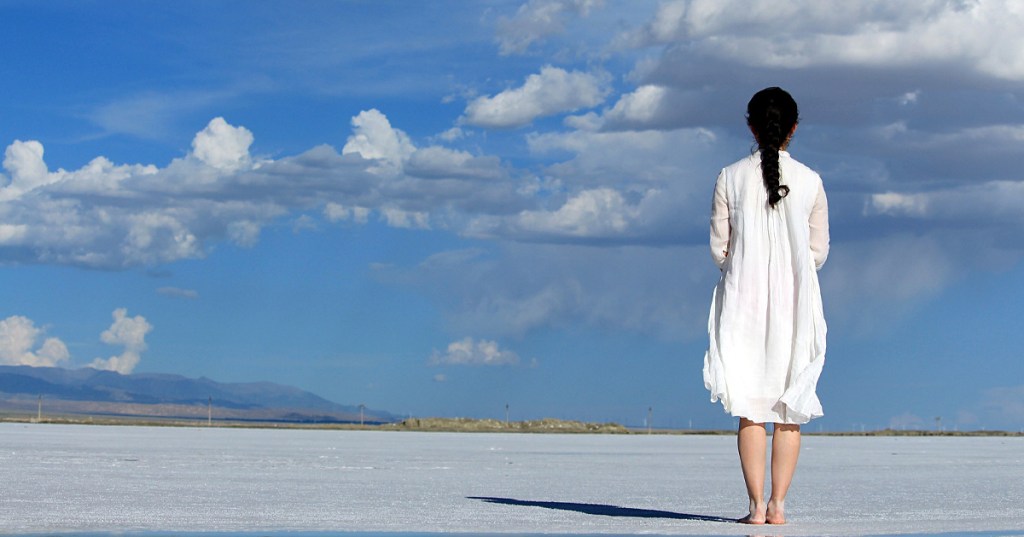 A woman standing on the sand, with beautiful blue skies, and looking back at what could have been.